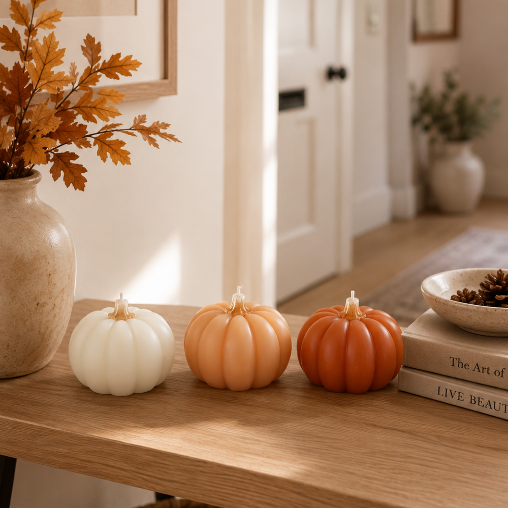 Three decorative faux pumpkins in white, peach, and terracotta orange are lined up on a wooden table in a warm, sunlit home interior.