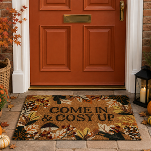 A rectangular coir doormat with a fall leaf pattern and the text 'COME IN & COSY UP' lies on a stone porch in front of a terracotta-colored front door. The scene is decorated for autumn with pumpkins, a lantern with a lit candle, and fall leaves.