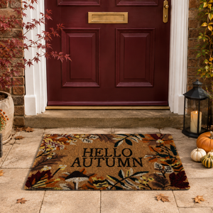 An autumn-themed doormat with the words 'HELLO AUTUMN' in black, featuring a colorful fall leaf and mushroom pattern in brown, beige, and orange tones, placed on a stone step in front of a red front door.