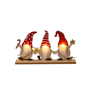 Three Christmas gnome figurines sitting side by side on a wooden base, each with a glowing warm light in its face. The gnomes have red hats, white beards, and are holding holiday items like a candy cane, a yellow bell, and a star, set against a plain white background.