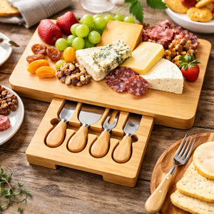A wooden cheese board set displayed on a rustic wooden table, featuring a rectangular wooden board topped with assorted cheeses, grapes, strawberries, nuts, and cured meats, with a matching drawer-style holder below holding four small cheese knives with wooden handles. A fork rests beside the board, and plates of food are partially visible around it.