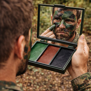 A person outdoors applies camouflage face paint from an open three-color makeup compact, with the mirror showing their face covered in green, black, and brown paint. The background is a blurred natural forest setting.