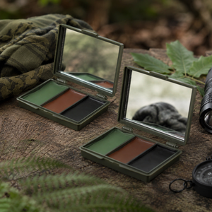 Two military-style compact powder makeup cases with mirrors, open on a rustic wooden surface outdoors. Each case contains three color pans: olive green, brown, and black.