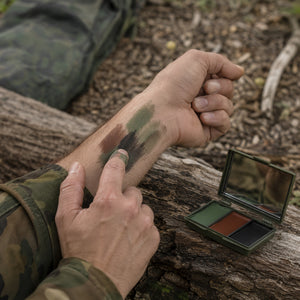 A close-up of a person outdoors applying camouflage makeup to their forearm, with a small open camouflage face palette resting on a log nearby.
