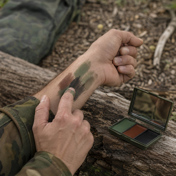 A close-up of a person outdoors applying camouflage makeup to their forearm, with a small open camouflage face palette resting on a log nearby.