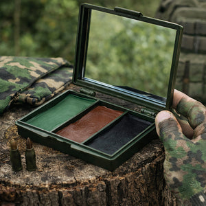 A camouflage military-style makeup case opened on a tree stump, showing three pans of color: green, brown, and black. A hand in a camouflage glove holds the case, with two bullets on the stump and a blurred outdoor background.