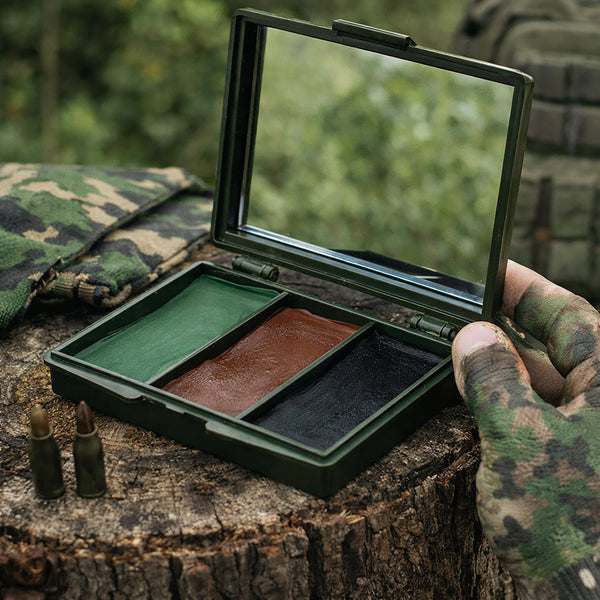 A camouflage military-style makeup case opened on a tree stump, showing three pans of color: green, brown, and black. A hand in a camouflage glove holds the case, with two bullets on the stump and a blurred outdoor background.