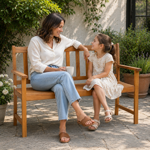 A woman and a young girl sit together on a wooden outdoor bench in a garden courtyard. The woman wears a white blouse, light blue jeans, and brown slide sandals, while the girl wears a white floral dress and white sandals.