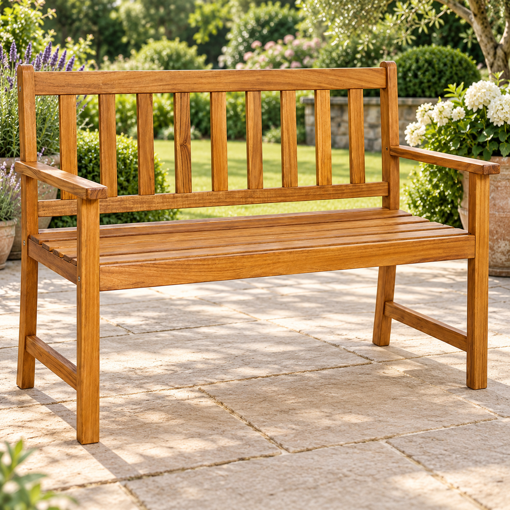 A wooden garden bench with a slatted backrest and armrests, shown outdoors on a stone patio with greenery and flowers in the background.
