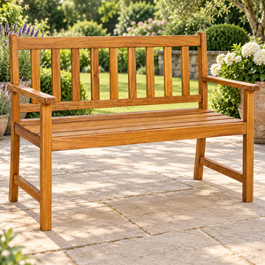 A wooden garden bench with a slatted backrest and armrests, shown outdoors on a stone patio with greenery and flowers in the background.