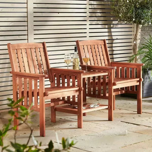 Two reddish-brown wooden outdoor armchairs with a matching small side table between them, set on a patio in front of a slatted wall with plants around.