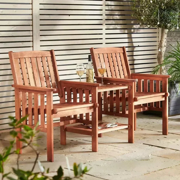 Two reddish-brown wooden outdoor armchairs with a matching small side table between them, set on a patio in front of a slatted wall with plants around.