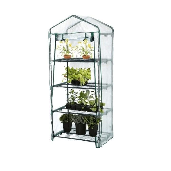 A four-tier portable greenhouse with a clear plastic cover and dark green metal frame, shown outdoors on a stone patio surrounded by garden plants and flowers.