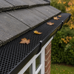 Black metal mesh gutter guard installed on a dark slate roof, shown with a few fallen autumn leaves resting on the mesh and gutter, with a brick house and garden in the background.