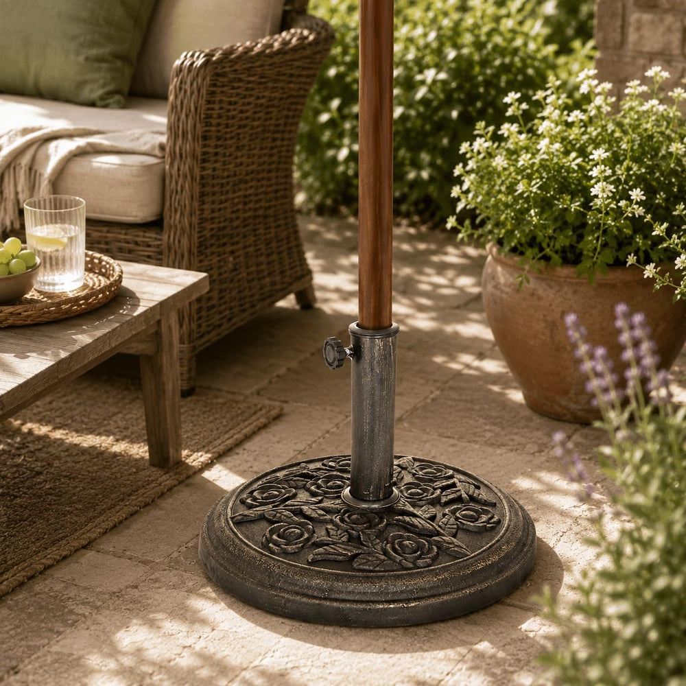 Outdoor patio umbrella base with a decorative cast-iron style round base and a vertical pole, shown in a garden patio setting next to a wicker sofa and potted plants.