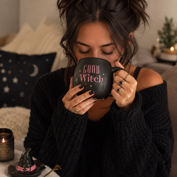 A woman in a black off-the-shoulder knit sweater holds a black ceramic mug with 'Good Witch' printed in pink lettering. The scene is set in a cozy, warmly lit bedroom or living space with a lit candle, a witch hat decoration, and pillows in the background.