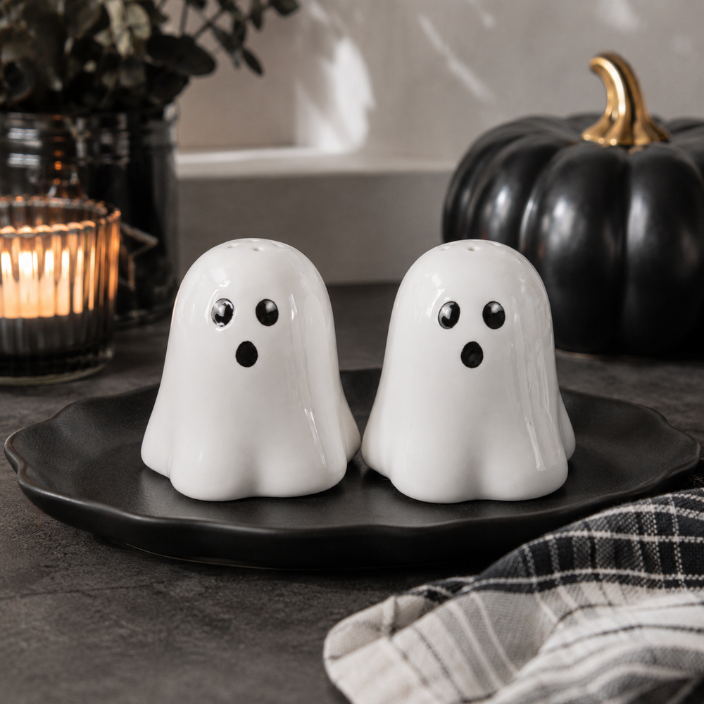 Two white ceramic ghost-shaped salt and pepper shakers with black eyes and mouths, sitting on a black scalloped platter. The scene is styled with a black decorative pumpkin, a lit candle, and a plaid cloth on a dark tabletop.