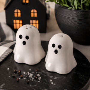 Two white ghost-shaped salt and pepper shakers with black eyes and mouths, sitting on a dark round tray with scattered peppercorns and coarse salt. A black house-shaped lantern and a potted plant are blurred in the background.