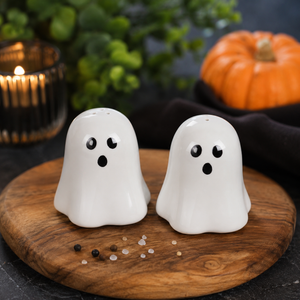 Two white ghost-shaped salt and pepper shakers with black eyes and mouths, displayed on a round wooden board with scattered peppercorns. The background features a dark cloth, green foliage, and a small orange pumpkin.