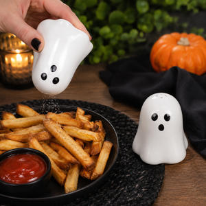 A hand holds a white ghost-shaped salt shaker above a plate of french fries with ketchup, with a matching ghost pepper shaker on a black cloth in the background.