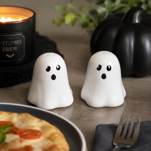 Two white ghost-shaped salt and pepper shakers with black eyes and mouths, placed on a tabletop with a candle, pumpkin, and food in the background.