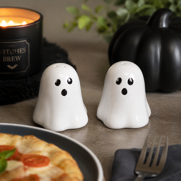 Two white ghost-shaped salt and pepper shakers with black eyes and mouths, placed on a tabletop with a candle, pumpkin, and food in the background.