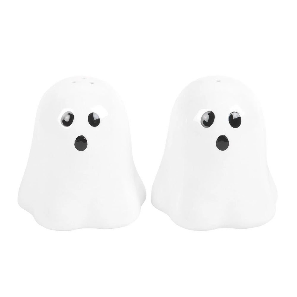 Two white ceramic ghost-shaped salt and pepper shakers with black eyes and mouths, sitting on a black scalloped platter. The scene is styled with a black decorative pumpkin, a lit candle, and a plaid cloth on a dark tabletop.