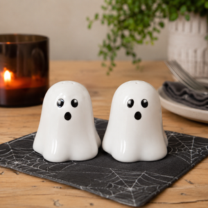 Two white ghost-shaped salt and pepper shakers with black eyes and mouths, sitting on a black spiderweb-patterned napkin on a wooden table with a blurred candle and plant in the background.