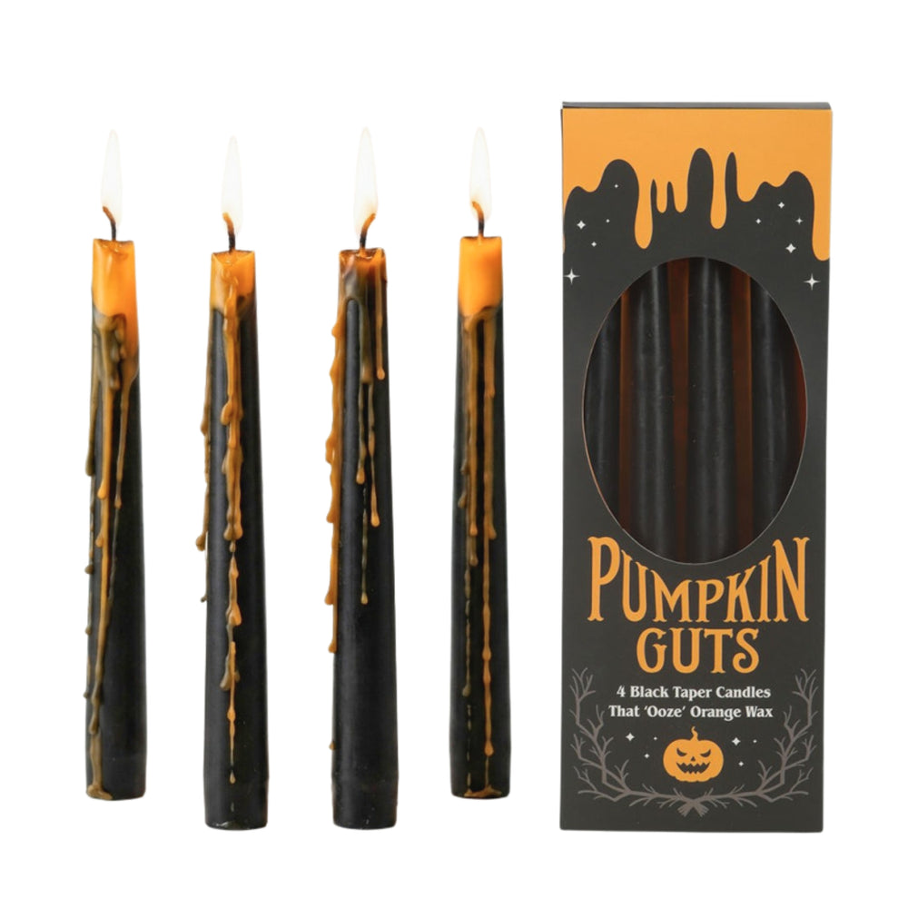 Four black taper candles with melted golden wax drips and lit flames, arranged on a wooden table in a warm, styled interior setting with books, a ceramic vase, and small food props.