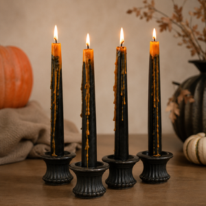 Four black taper candles with orange wax drips and lit flames, displayed in matching black holders on a wooden surface with a warm autumnal background.