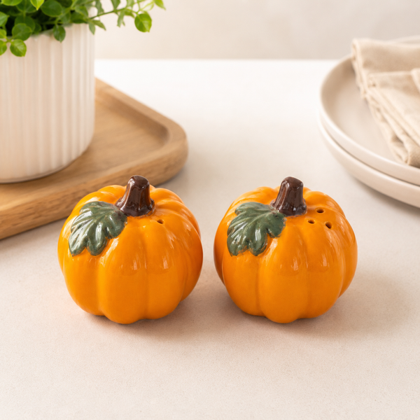Two small orange pumpkin-shaped salt and pepper shakers with green leaves and dark brown stems, placed on a light tabletop with a blurred plant and plates in the background.