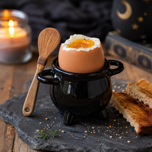 A small black ceramic cauldron-shaped egg holder with a halved boiled egg inside, styled on a dark stone plate with a wooden spoon, toast, and a blurred candle in the background.