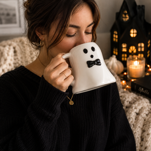 A woman in a black ribbed sweater holds a white ceramic mug shaped like a ghost with a black face and bow tie, set in a cozy, warmly lit room with a lit candle and a black haunted house decoration in the background.
