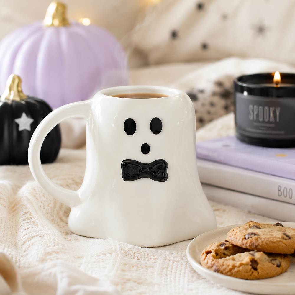 A white ceramic mug shaped like a ghost with a black face and bow tie, filled with coffee and styled on a cream knit blanket with cookies, pumpkins, a candle, and books in the background.