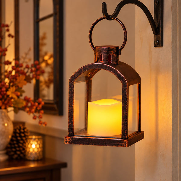 A rustic dark bronze lantern-style wall sconce with a glowing warm yellow LED candle inside, mounted on a beige wall in a cozy interior setting.