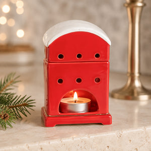 A red ceramic holiday candle warmer with a white snow-dusted roof, holding a lit tealight candle, set on a light stone surface with blurred holiday decor in the background.