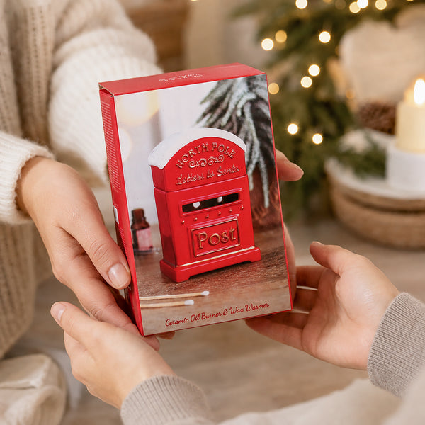 A person holds a red gift box featuring an illustration of a red Christmas North Pole Santa mail box. The scene is set in a cozy indoor environment with warm holiday lights and a blurred Christmas tree in the background.