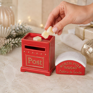 A hand places cream-colored wax melts into a red and gold North Pole post box, with a matching red and white 'North Pole Letters to Santa' container beside it on a festive tabletop.