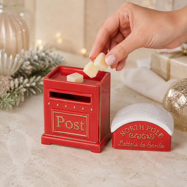 A hand places cream-colored wax melts into a red and gold North Pole post box, with a matching red and white 'North Pole Letters to Santa' container beside it on a festive tabletop.