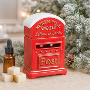 A red North Pole Letters to Santa post box candle holder with a white snow-dusted top, styled on a wooden surface with a small amber dropper bottle and cream wax cubes, with blurred festive greenery in the background.
