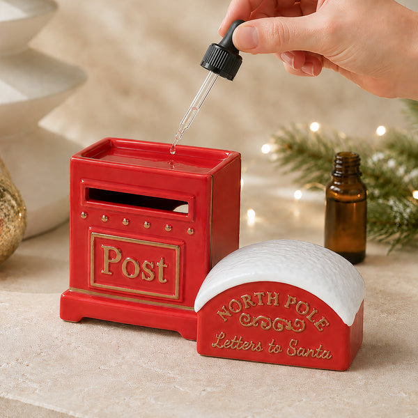 A red ceramic post box and a matching North Pole letters to Santa holder are shown on a beige surface with a hand holding a dropper above the post box, plus a small amber bottle and soft holiday lights in the background.