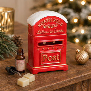 A red ceramic North Pole letters-to-Santa post box with gold lettering and a white faux-snow top, displayed on a wooden table with holiday decor including a small bottle, dropper, soap bars, pine branches, and gold ornaments against a blurred Christmas tree background.