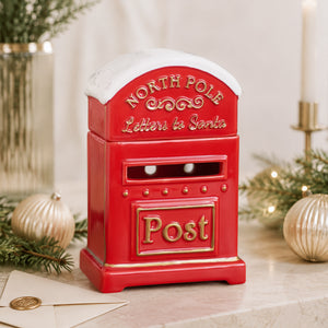 A red ceramic North Pole post box with gold lettering and a white faux-snow top, styled on a tabletop with holiday greenery, gold ornaments, and a candle in the background.
