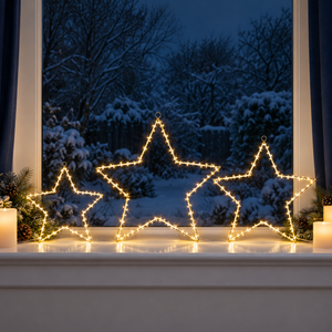 Three large star-shaped Christmas string lights glowing warm yellow on a white windowsill, with a snowy outdoor scene visible in the background and candles on both sides.
