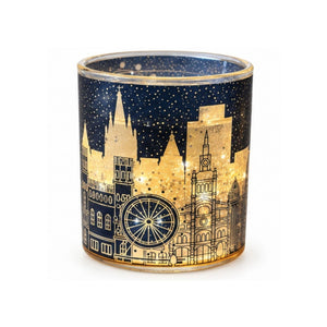 A cylindrical glass candle holder featuring a gold glittery skyline design with spires, a Ferris wheel, and stars on a navy blue background, shown lit from within against a white background.