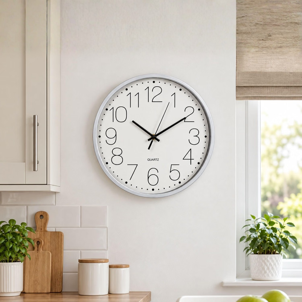 A round wall clock with a white face, black numerals and hands, and a silver-toned outer rim, mounted on a light beige wall in a bright kitchen setting.