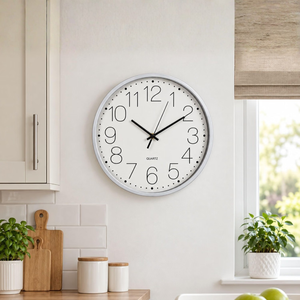 A round wall clock with a white face, black numerals and hands, and a silver-toned outer rim, mounted on a light beige wall in a bright kitchen setting.