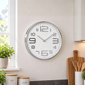 A round wall clock with a silver frame and white face, featuring large black Arabic numerals and silver hands, mounted on a beige wall in a bright kitchen setting.
