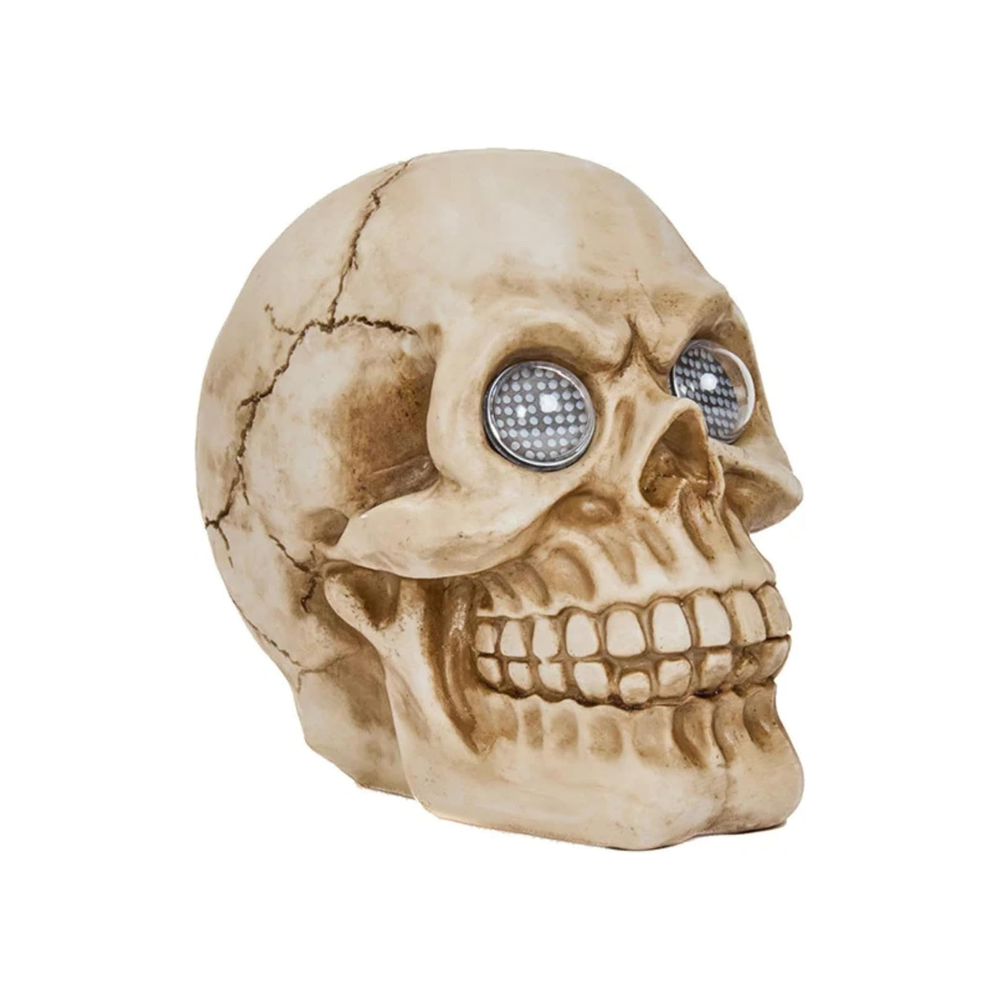 A decorative skull garden statue with glowing blue LED eyes, displayed on a stone base outdoors among green foliage and white gravel.