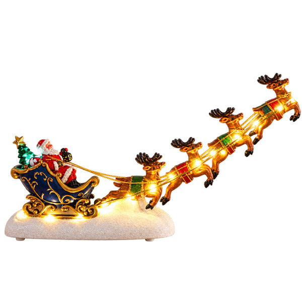 A Santa Claus Christmas ornament figurine in a flying sleigh pulled by four reindeer, set on a glittery white base against a plain white background. The sleigh is dark blue with gold trim, Santa is dressed in red and white, and the reindeer are gold with red and green accents.